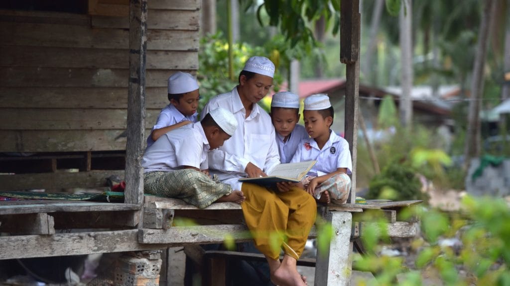 man surrounded by 4 young boys reading from a book