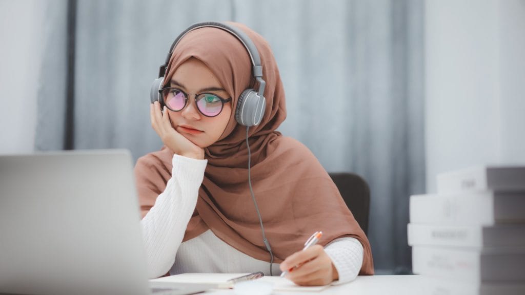 girl with hijab looking into a laptop, with pen in hand and headphones on head